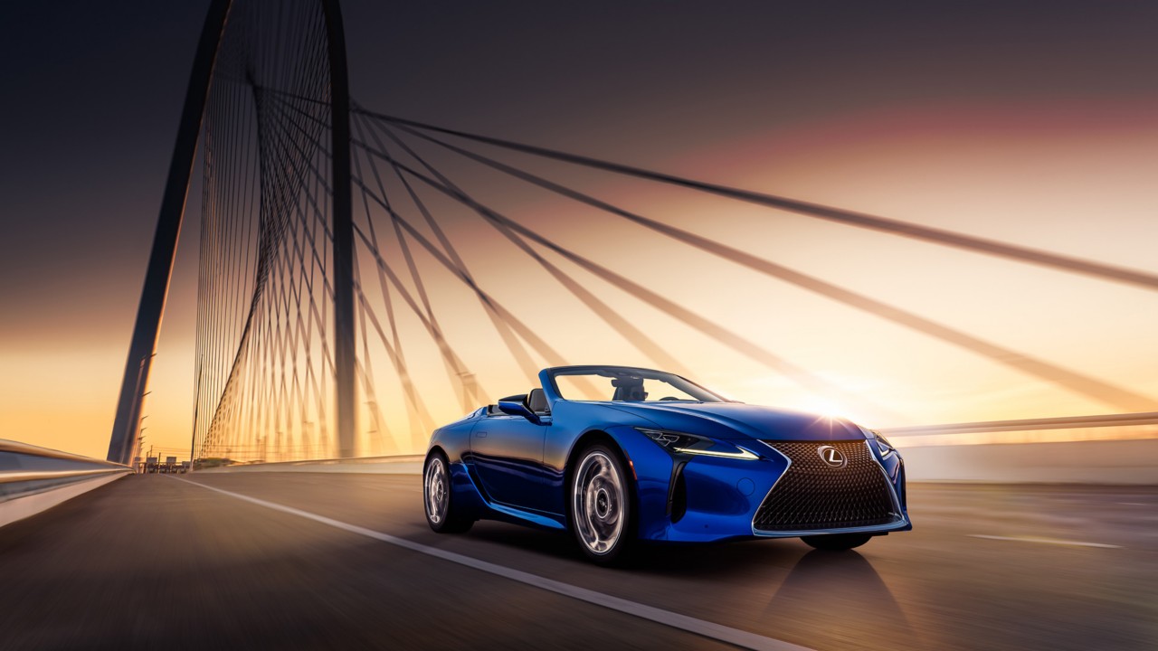 Ein blaues Lexus Cabrio fährt bei Sonnenaufgang über eine moderne Brücke.